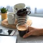 Person holding a full glass of coffee on a background of multiple empty finished cups.