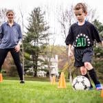 Kid kicking a soccer ball while his private coach observes.