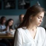 A sad young woman avoiding a group peers and sitting alone in a cafe.