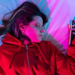 A young girl looking at her smartphone and doom-scrolling on the bed in her neon-lit room.