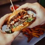 A bacon burger being held with a plate of fries in the background.