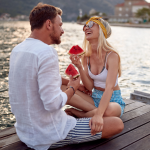 A couple having good time on a date, sitting on a wooden dock, laughing and eating watermelon slices.