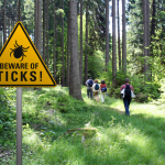 Warning sign near a lush green forest trail that says "beware of ticks" with hikers in the background