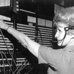 Female CDC telephone operator seated at a switchboard wearing a traditional headset, reaching for a telephone patch cable, 1974.