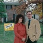 Portrait of couple standing next to For Sale sign in front of house
