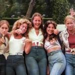 Color photo of five teenage girls leaning against a car on a tree-lined suburban street in San Diego in 1977. They wear high-waisted jeans and various casual tops including a tied blouse, a floral shirt, and a cardigan. One makes a peace sign and another rests her head on a friend's shoulder. Green hedges and parked cars are visible behind them.