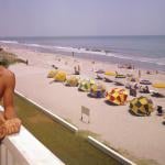 A smiling couple lean on a balcony at Thunderbird Motel, which overlooks a section of beach shaded by various colored umbrellas, Myrtle Beach, South Carolina, 1960s.