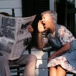 Senior couple sitting on a porch in rocking chairs. Husband is reading the paper and the wife is shouting at him. 