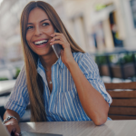 A young woman with long hair and a big smile sitting at the table outdoors holding a cellphone to her ear.