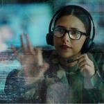 A woman wearing military camouflage and glasses, along with a headset. She is looking on the digital display in front of her.