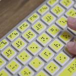 A yellow computer keyboard with braille and someone's fingers typing.