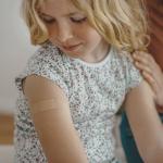 A young child with a small adhesive bandage on their upper arm. The child is wearing a floral-patterned shirt and looking down at the bandage.