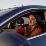 A close up of a young woman sitting in the driver’s seat of a car, smiling warmly.