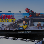 A billboard advertisement promoting the upcoming movie Deadpool & Wolverine by Marvel Studios, set to release on July 26. There is a man dressed in black and red bodysuit lying on top of a damaged car, while a man is in the driver's seat looking focused and intense.