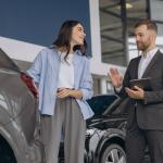 A man dressed in a dark suit holding a tablet is looking towards a young woman in front of him. The woman is casually dressed in a light blue button-down shirt over a white top and gray trousers. She is smiling and appears to be listening to the man next to her. Both individuals are standing next to a car. 