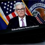 A close-up of a video or broadcast of a "Federal Reserve" news conference on a laptop screen. A man with gray hair is wearing glasses dressed in a suit with a white shirt and purple tie.