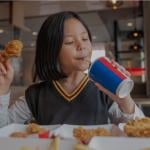 A girl sipping a drink and holding a piece of fried chicken sitting in a restaurant. 