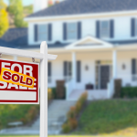 A for sale sign with a sold sticker on it in the front lawn of a midsize suburban home.