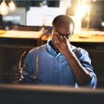 A tired man in a dimly lit workspace sitting at a desk.