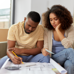 A concerned young couple reviewing their finances while looking at a blueprint for their home. 