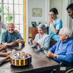 A group of older adults play games at a table with medical staff smiling behind them.