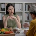 A woman looking at the older woman sitting in front of her. They are in the office with files in the background and a plate of vegetables and fruit on the desk.