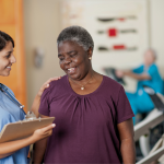 An older Black woman standing close to the nurse with a clipboard.