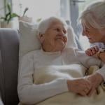 An elderly woman with white hair reclining in a chair, covered with a soft beige blanket. She is wearing a white sweater and a pearl necklace. Beside her, a middle-aged woman leans in holding the elderly woman's hand and smiling.