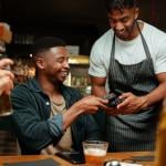 African American man is holdiong his phone close to a credit card reader held by a waiter at a table.