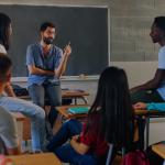 A group of students seated around a teacher in a classroom setting. The teacher casually dressed, is sitting on a desk and appears to be engaged in a discussion with the students. 