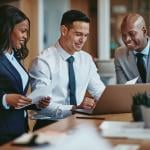 Three smiling businesspeople looking at the laptop on a table in an office.