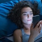 Young woman lies in bed in a dark room while looking at her phone.