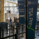 Two blue signs with white lettering suspended on a pole at an airport. Large one says "Transportation Security Administration TSA Pre" and the smaller one says 'Transportation Security Administration enter here.'