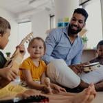 A young male teacher sitting on the floor with a tambourine in his hand surrounded by young kids.