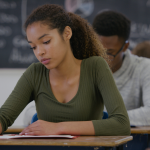 African American teenage girl sitting at the desk in a classroom holding a pencil and looking down at the paper in front of her.  