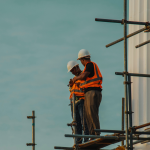 Two men in bright-orange vests wearing hard hats are standing on scaffoldings holding tools.