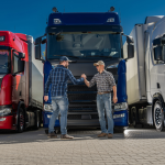 Two men in checked shirts, jeans, and baseball hats shaking hands. There are three truck cabins in the background behind them. 