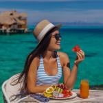 A youthful woman in a straw hat lounges al fresco at a table beside blue waters, delicately grasping a slice of watermelon, with a platter of fresh, vibrant fruits in the the foreground.