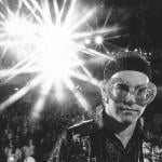 Black and white image of Elton John in bright lights wearing big sunglasses.