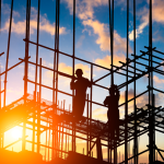 Workers stand on scaffolding in a construction site with an orange sunset in the background.