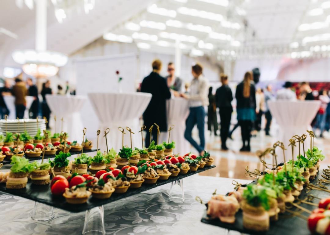 Trays of catered food with people at an event in the background.