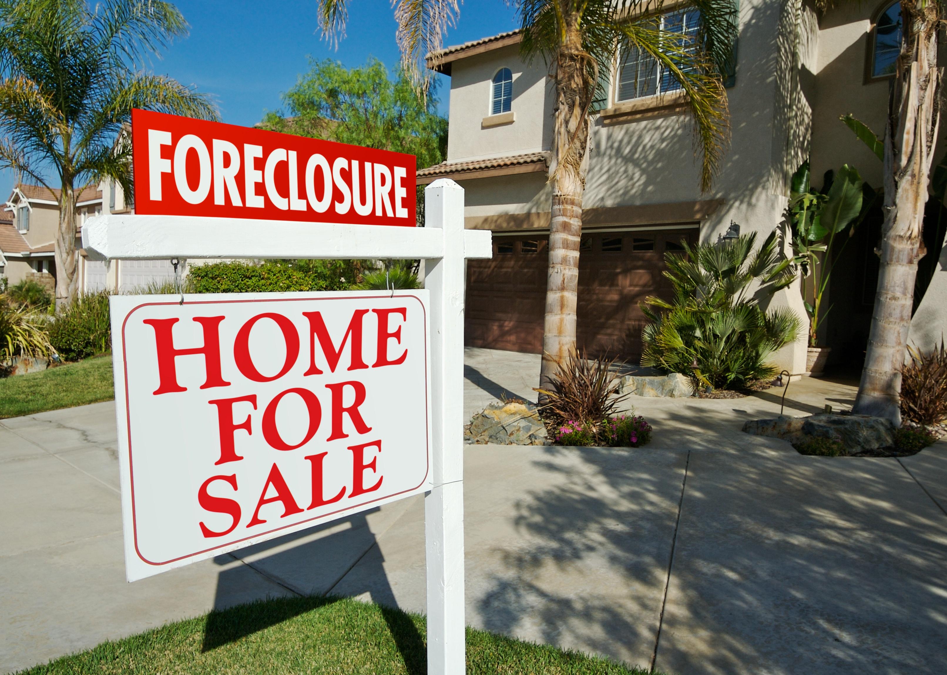 A brown stucco home with palm trees and a foreclosure sign in the front yard.