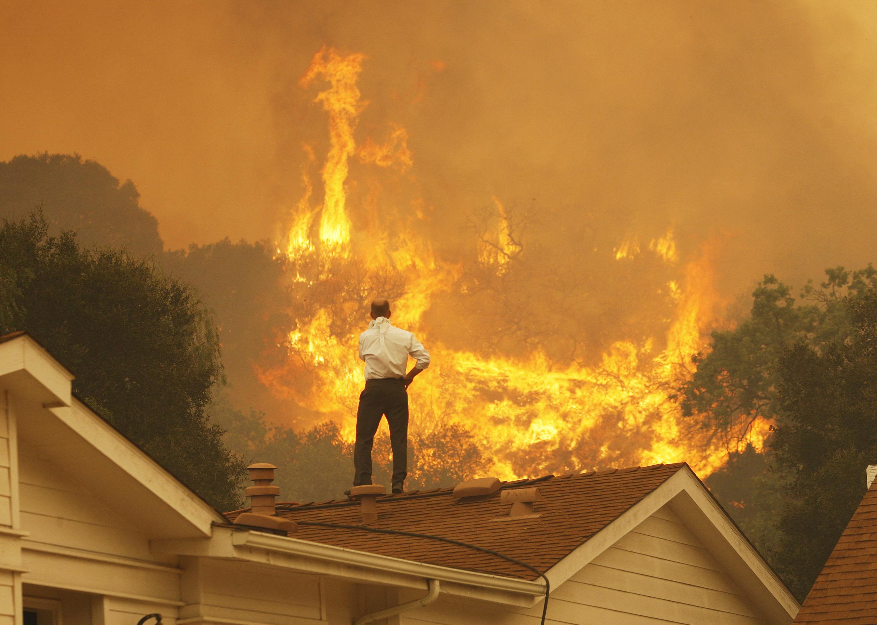 A man on a rooftop looks at approaching flames.