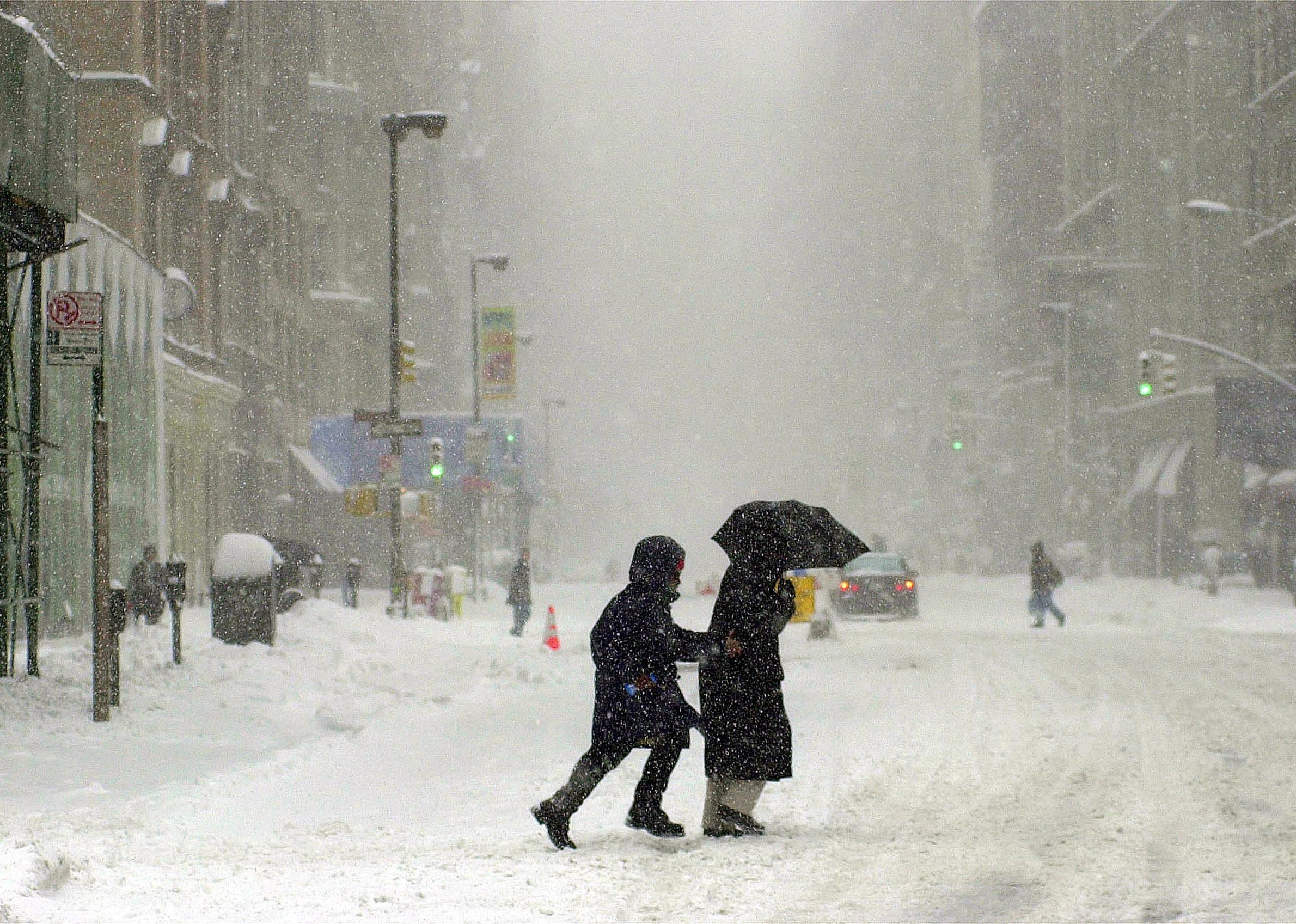 Two people make their way across a street with snow falling around.
