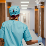 A worker in green scrubs walks down a hospital hallway pushing an IV bag.
