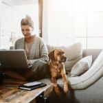 Woman working on laptop while sitting on couch with a dog.