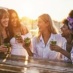 A group of women laughing and drinking cocktails outside as the sun is setting. 