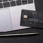 Black premium bank card lays on a silver macbook laptop computer next to the trackpad, with a shiny fancy pen on the table.