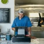 Person wearing a blue shirt entering items into a tablet cash register behind the counter with a kitchen and sign int he background.