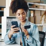 Black woman entrepreneur looking at a business credit card and holding smartphone inside an office with boxes and shelves and a blurred blond person in the background. 